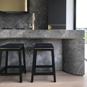In front of grey granite kitchen island bench sits two kitchen stools finished in solid wood stained black with a soft wool seat in grey