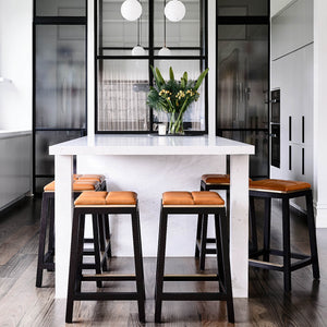 Sunlight drips in through large kitchen windows highlighting a gorgeous large white stone kitchen island with black wood stools clustered around three sides featuring brass footrests and tan leather seats 